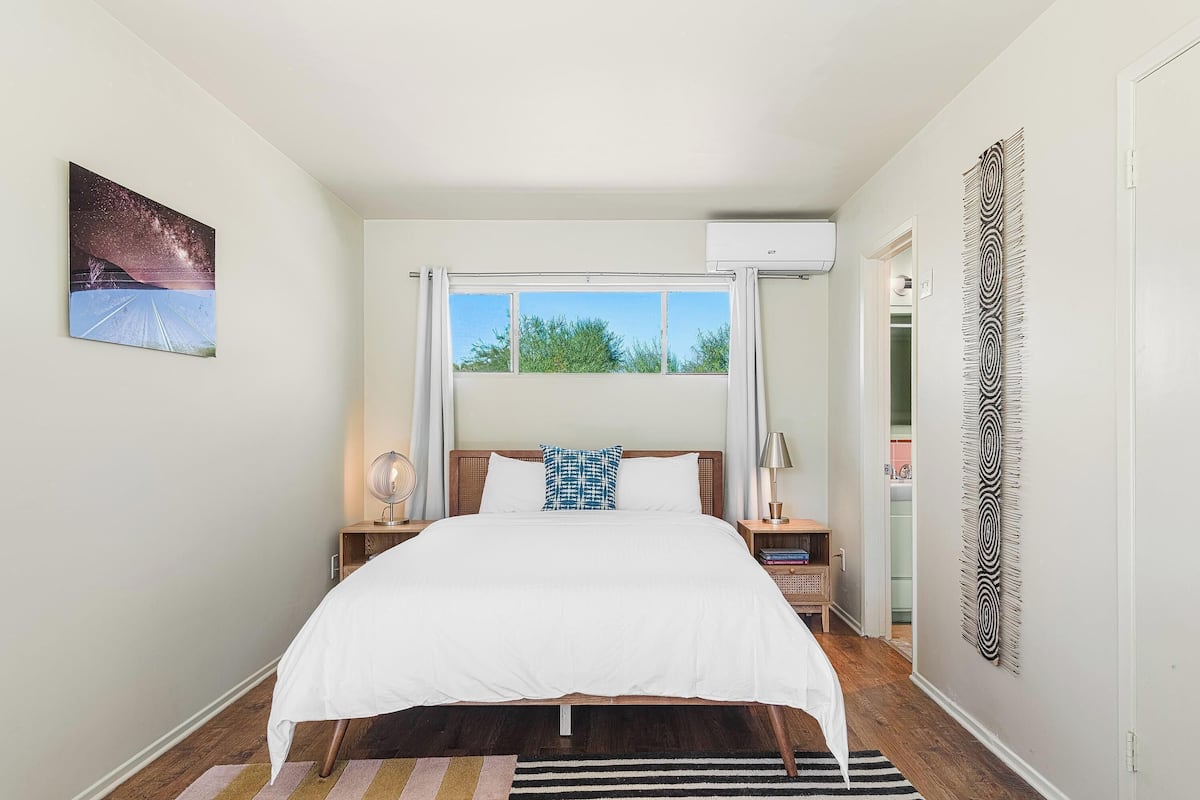 Une chambre à coucher comprenant un lit, une fenêtre, un climatiseur fixé au mur, une table de chevet avec une lampe et un élément décoratif mural.