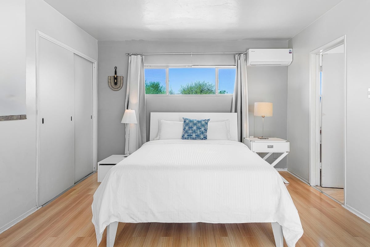 Une chambre à coucher comprenant un lit, une table de chevet, une lampe, un appareil de climatisation et une fenêtre donnant sur un paysage verdoyant.