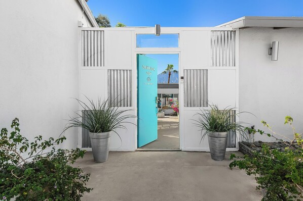 A white building with a turquoise door labeled 'Nurturing Nest.' - Nurturing Nest (Desert Hot Springs)