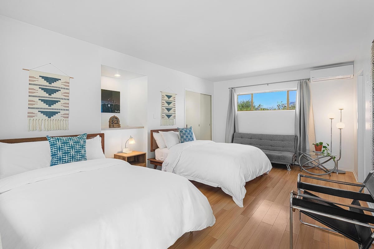 Une chambre d’hôtel avec deux lits, un bureau, une chaise, une fenêtre donnant sur un beau paysage et une plante.