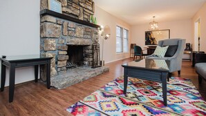 Una sala de estar con chimenea de piedra, una alfombra colorida y un sillón gris.
