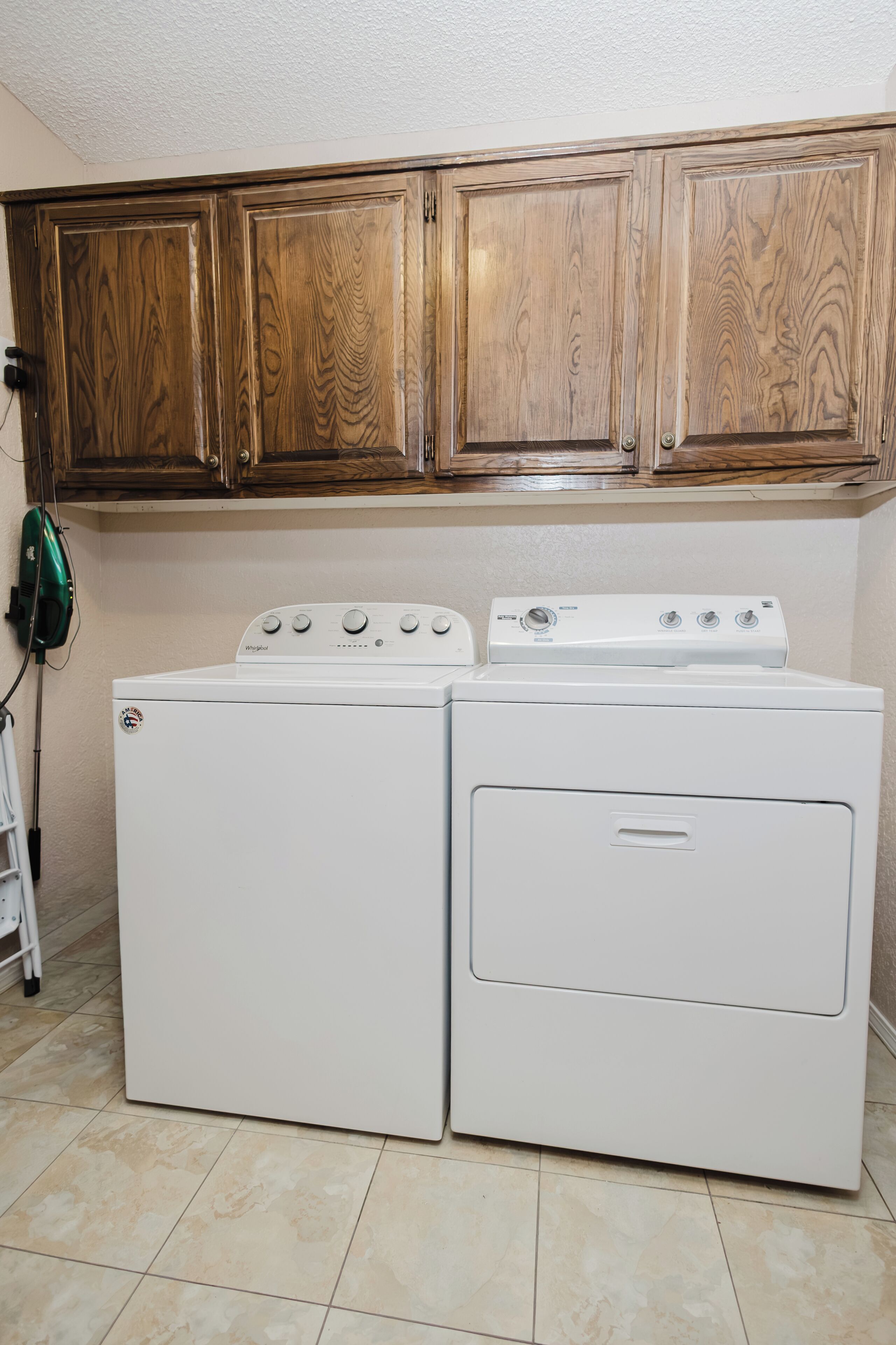 Une buanderie équipée de deux lave-linge et deux sèche-linge à chargement frontal, tous deux de couleur blanche, avec des armoires en bois au-dessus et un câble d’alimentation vert fixé au mur.