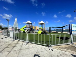 Ein Spielplatz mit Rutschen und Klettergerüsten, umgeben von einem Metallzaun, unter blauem Himmel.