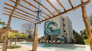 A pool area with a wooden pergola, lounge chairs, and a building with a wave mural.