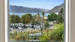 Vista desde una ventana sobre un estacionamiento con coches, árboles y un lago con montañas al fondo.