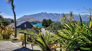 Vista de un paisaje montañoso con una masa de agua en la distancia, un edificio moderno con techo oscuro, y diversas plantas en primer plano.