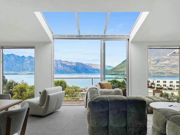 A modern living room with a large skylight, comfortable seating, and a view of mountains and a lake.