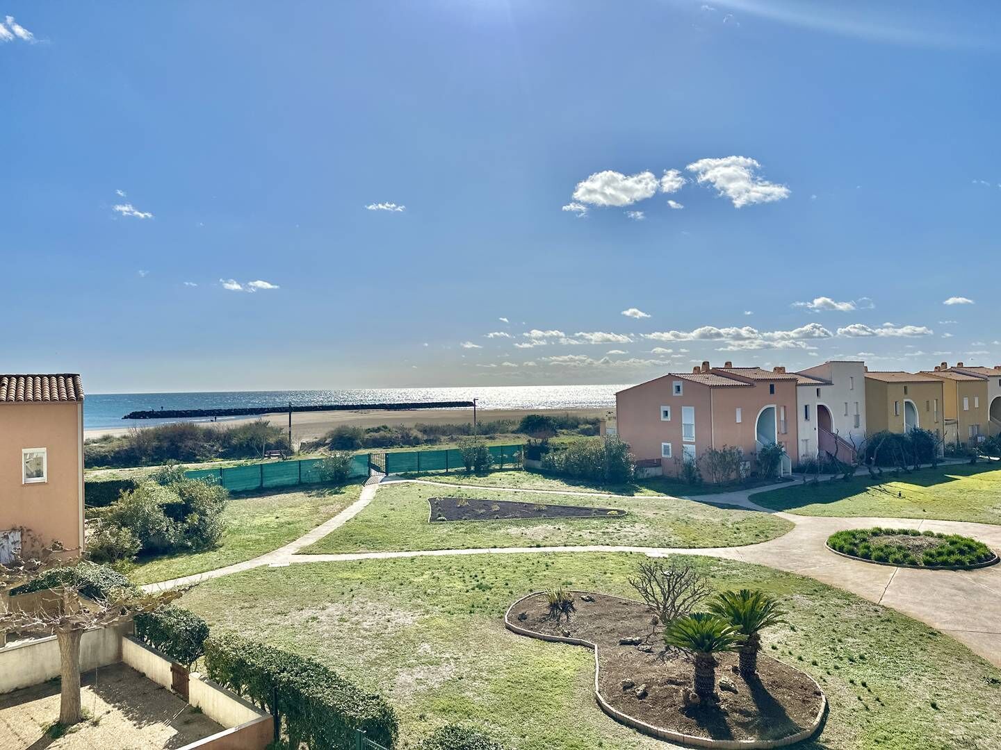A coastal residential area with a clear view of the sea, a paved walkway, and a well-maintained lawn with a circular garden.