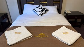 A neatly made bed with a white duvet, a black and white pillow, and two folded towels on a brown bedspread with the logo "CAMINO REAL". - Camino real (Puno)