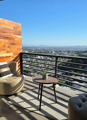 A balcony with a wooden wall, a cushioned chair, a small table with a plant, and a view of a cityscape.