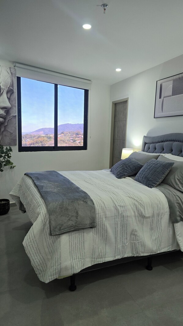 A bedroom with a large window, a bed with grey bedding, and a view of a mountainous landscape.