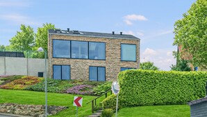 Une maison moderne à deux étages, dotée d’une façade en pierre, de grandes fenêtres et d’un jardin bien entretenu.