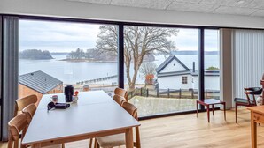 Un espace repas avec une grande table et des chaises, une vue sur un lac et une maison située de l’autre côté de l’eau.