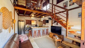 A rustic interior with a wooden staircase, a kitchen area, a seating area with a sofa and stools, and a television. - Pousada Reserva Setiba (Guarapari)