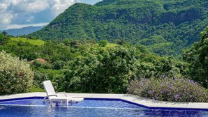 A pool with a white lounge chair, surrounded by lush greenery and a mountain backdrop.