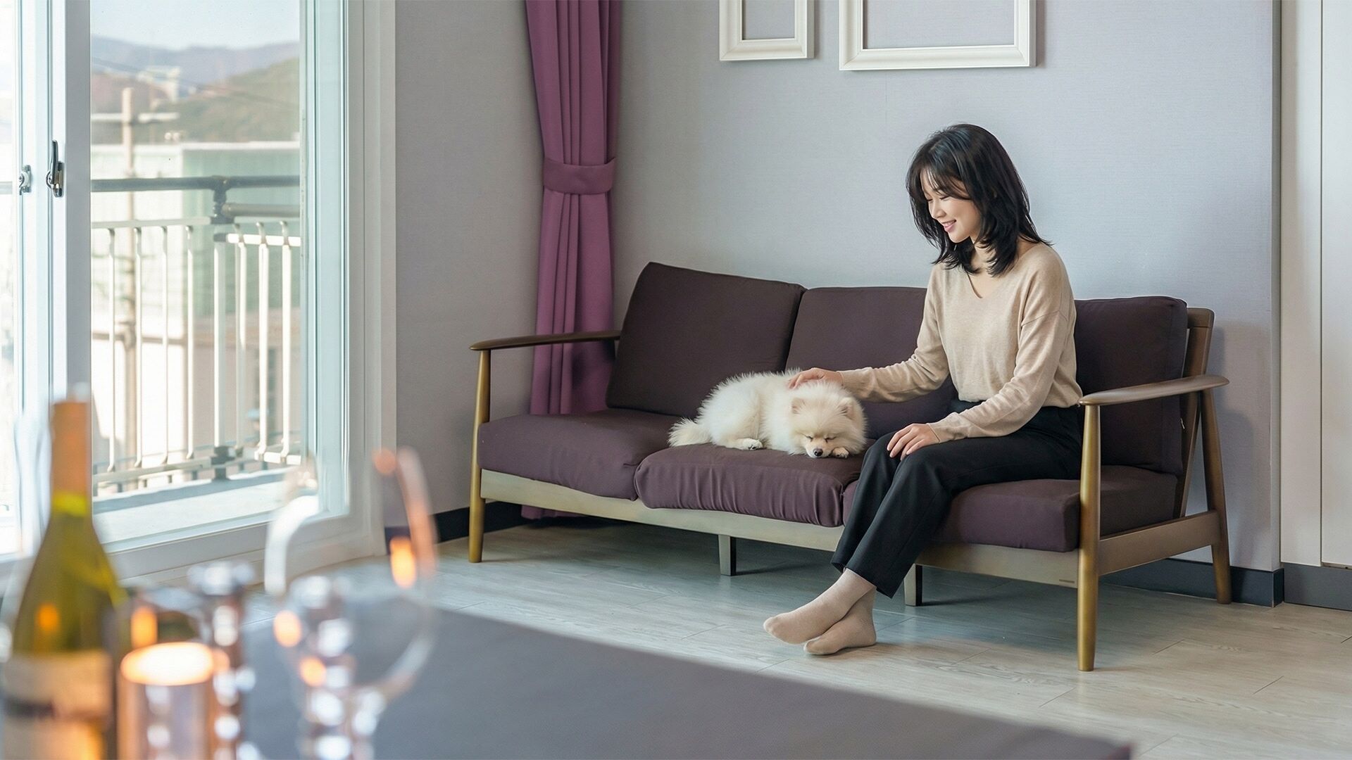 A person sitting on a sofa with a fluffy dog, a window with curtains, and a table with candles.