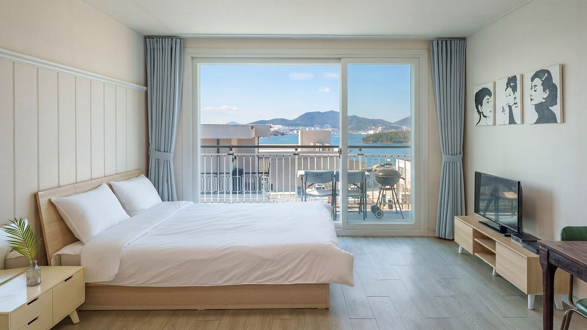 A hotel room with a bed, a TV, a desk, and a balcony with a view of the sea and mountains.