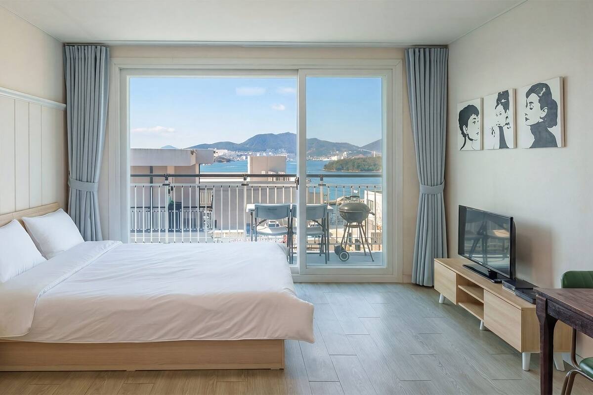 A hotel room with a bed, a TV, a desk, and a balcony with a view of the sea and mountains.