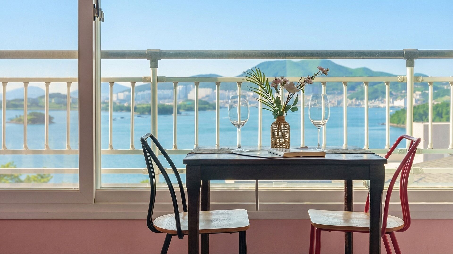 A balcony with a table, two chairs, a vase with flowers, and a view of the sea and mountains.