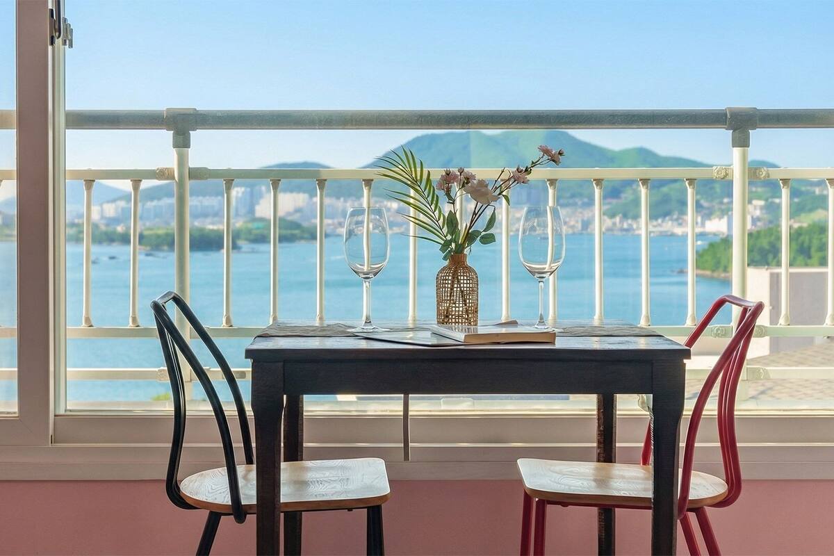 A balcony with a table, two chairs, a vase with flowers, and a view of the sea and mountains.