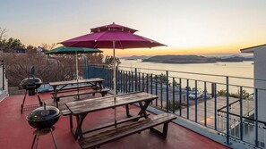 A balcony with picnic tables, a grill, and umbrellas overlooking a body of water. - Yeosu Blooming Ocean View Family Pension (Yeosu)