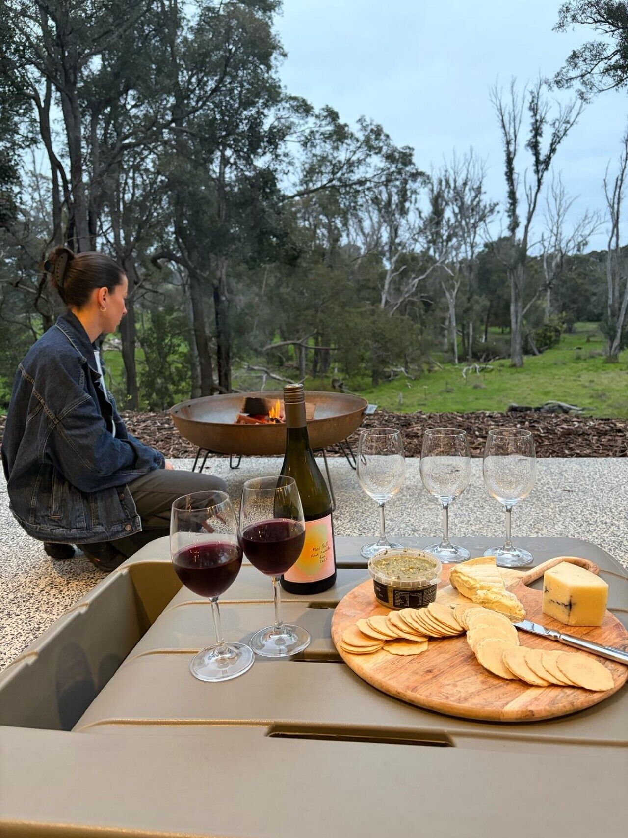 An outdoor dining setup with a table, wine glasses, a bottle, and cheese.