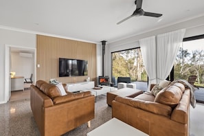 A modern living room with leather sofas, a flat-screen TV, a fireplace, and a large window with curtains. - Nannup Bush Retreat · Firepit, Walk to Town (Nannup)