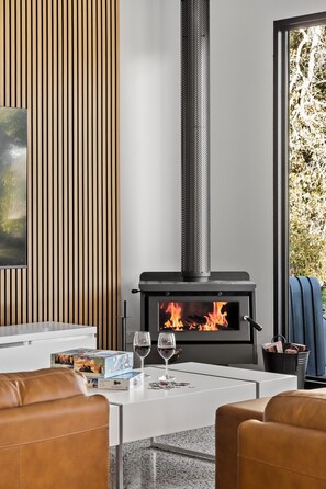 Un salon moderne avec un poêle à bois, une table basse blanche, des fauteuils en cuir et une vue par la fenêtre.
