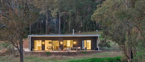 Una casa moderna con un amplio ventanal, un patio con mobiliario exterior y un fondo de bosque.