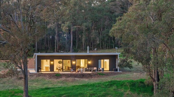 A modern house with a large glass door, a patio with outdoor furniture, and a forested background. - Nannup Bush Retreat · Firepit, Walk to Town (Nannup)
