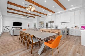 A modern kitchen with white cabinets, a central island, and a dining area with wooden chairs and a long table. - Beachfront + Hot Tub | Game Room + Pets | Sleep 18 (Galveston)