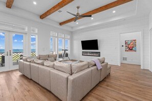 A spacious living room with a large beige sectional sofa, a flat-screen TV mounted on the wall, and a balcony with a view of the sea. - Beachfront + Hot Tub | Game Room + Pets | Sleep 18 (Galveston)