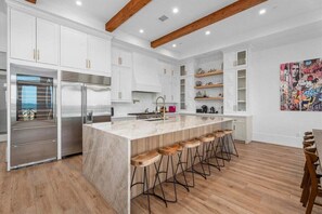 A modern kitchen with white cabinetry, a large island with bar stools, stainless steel appliances, and a wooden ceiling beam. - Beachfront + Hot Tub | Game Room + Pets | Sleep 18 (Galveston)