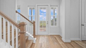Un vaste hall d’entrée avec un escalier en bois, des murs blancs et une porte donnant sur un balcon d’où l’on a une vue dégagée sur le ciel.