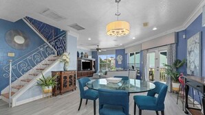 A spacious living area with a glass-top dining table, blue upholstered chairs, a staircase with decorative railings, and a large chandelier.