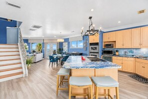 A modern kitchen with a central island, wooden cabinets, and a dining area with blue chairs. - Galveston Canal Home | Large Deck | Sleeps 14 (Galveston)