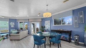 A modern living room with a glass dining table, blue chairs, a beige sofa, and a large window.