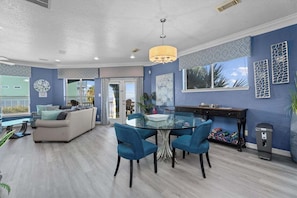 A modern living room with a glass dining table, blue chairs, a beige sofa, and a large window. - Galveston Canal Home | Large Deck | Sleeps 14 (Galveston)