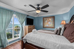 A bedroom with a bed, a ceiling fan, a TV, a painting, and a view of the ocean. - Galveston Canal Home | Large Deck | Sleeps 14 (Galveston)