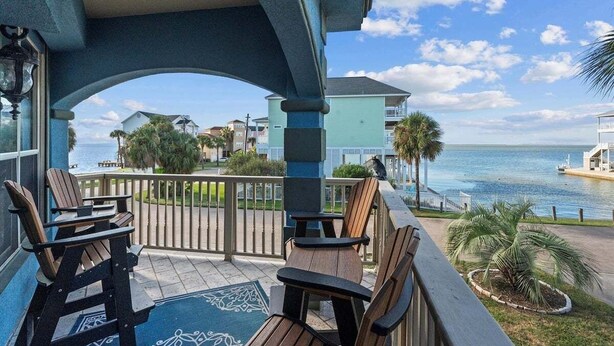 A balcony with wooden chairs and a patterned rug, overlooking a marina and buildings by the water.