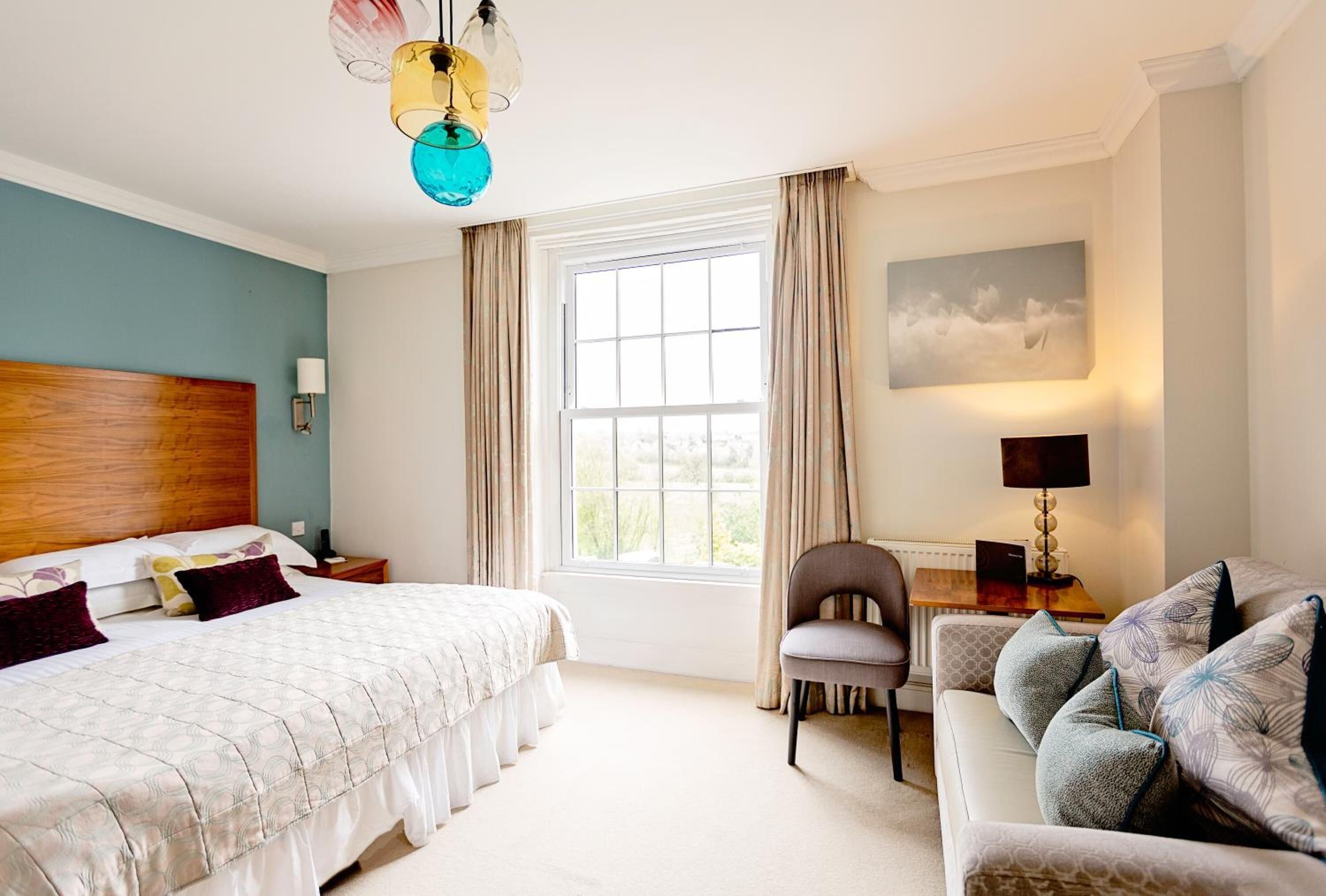 A hotel room with a large bed, a desk, a chair, and a couch. There is a window with curtains, a painting on the wall, and a lamp on the desk.