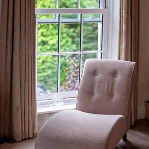 Une chaise près d’une fenêtre avec des rideaux, un vase sur le rebord de la fenêtre et une vue sur la verdure à l’extérieur.