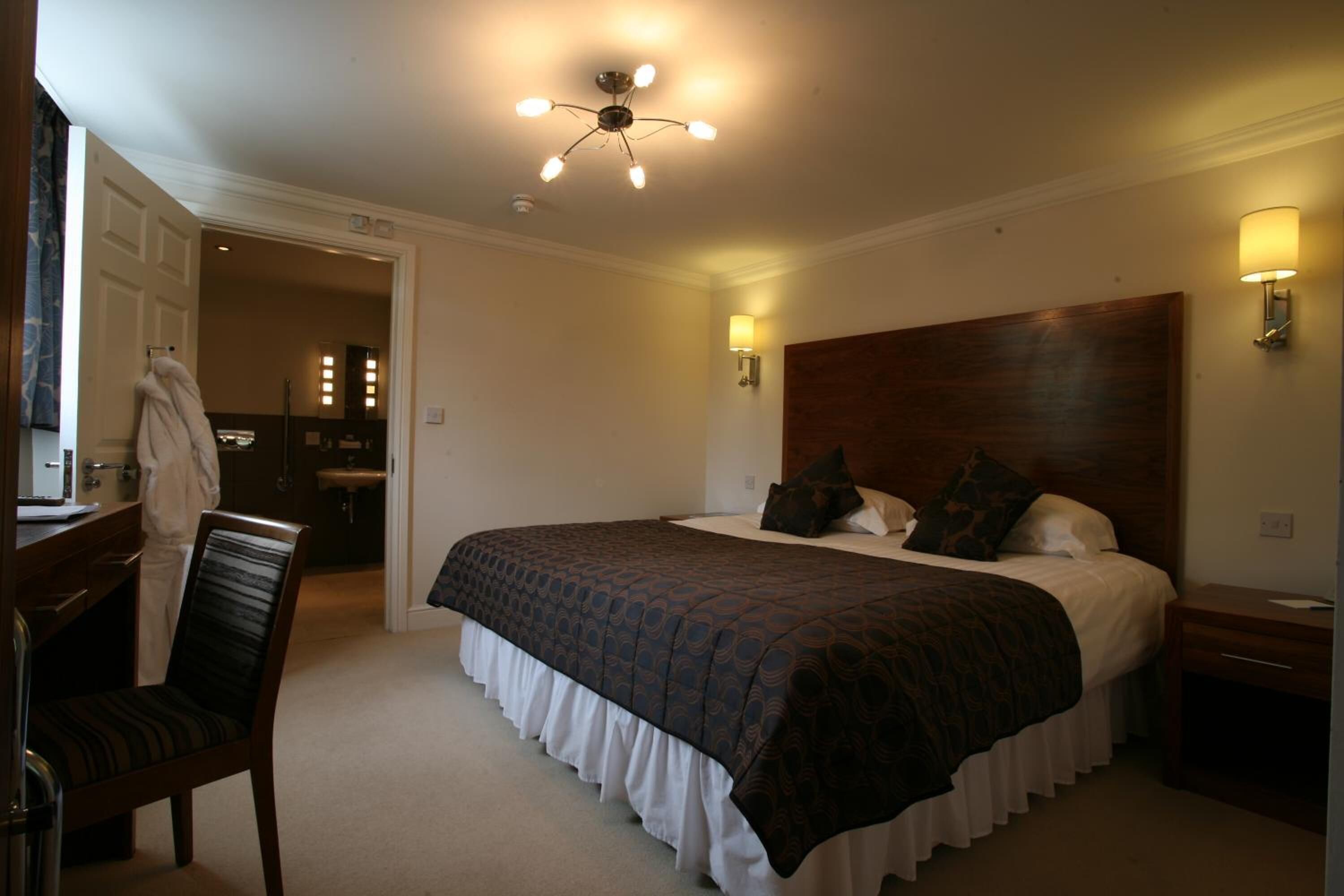 Hotelkamer met een groot bed, een houten hoofdbord, een bureau met stoel, en een zichtbare badkamer door een open deur.