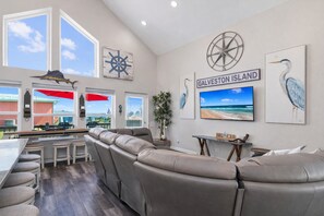 Een ruime woonkamer met een grote leren bank, een keuken met een bar, en een televisie waarop een strandscène te zien is.