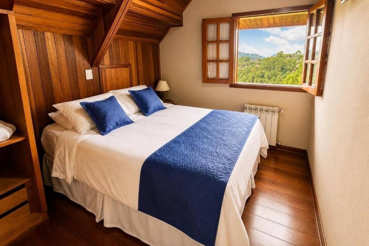 Ein Schlafzimmer mit Holzbalkendecke, einem großen Bett mit weißblauer Bettwäsche, einem Fenster mit Blick ins Grüne und einer Heizung.