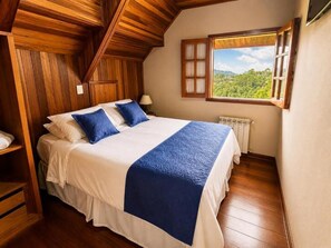 A bedroom with a wooden ceiling, a large bed with white and blue bedding, a window with a view of greenery, and a radiator. - Pousada Chateau Elegance (Campos do Jordão)