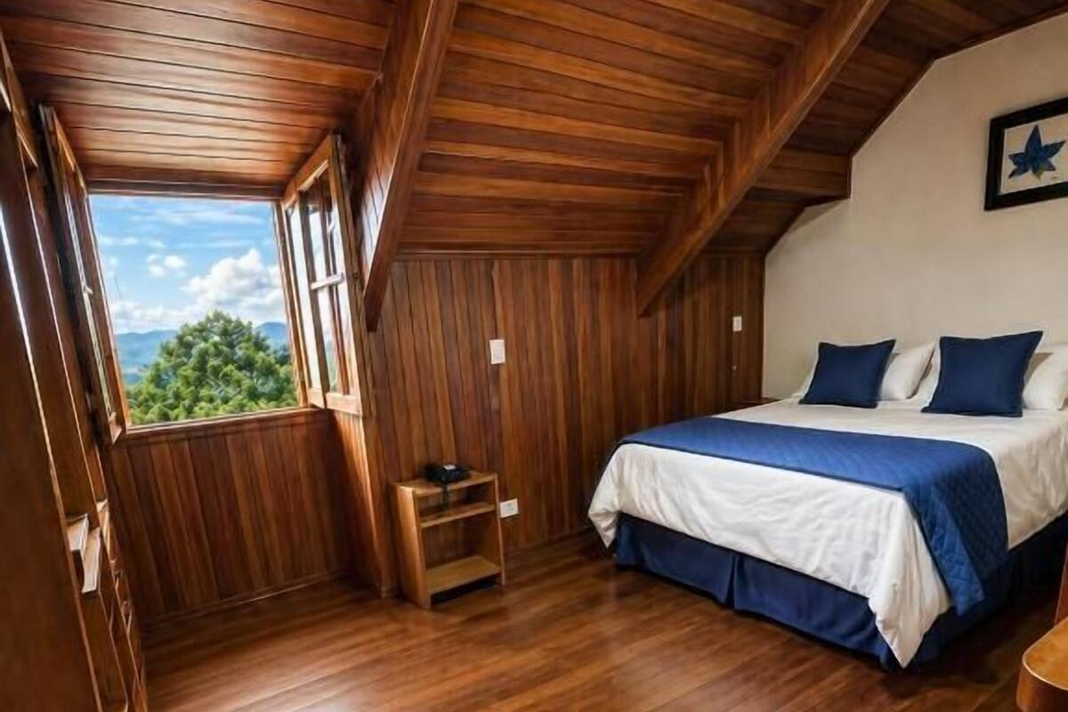 Ein Schlafzimmer mit Holzboden, einem Bett mit blauer Bettwäsche, einer Holzzieldecke und einem Fenster mit Blick auf Bäume.
