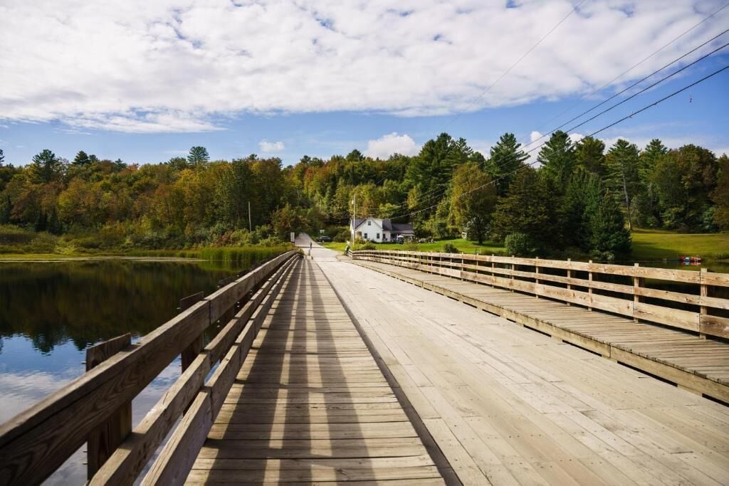 Un pont en bois enjambe un lac paisible, avec une maison qui se profile à l’arrière-plan.