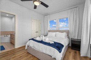 A bedroom with a bed, a ceiling fan, a window with curtains, a nightstand, and a view of the sky. - Beachfront | Wraparound Deck | Steps to Sand (Bolivar Peninsula)