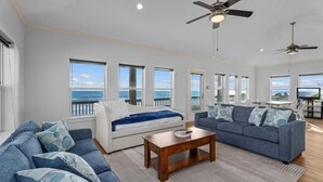 A spacious living room with a large sofa, a smaller sofa, a coffee table, and a TV. There are multiple windows offering a view of the ocean.